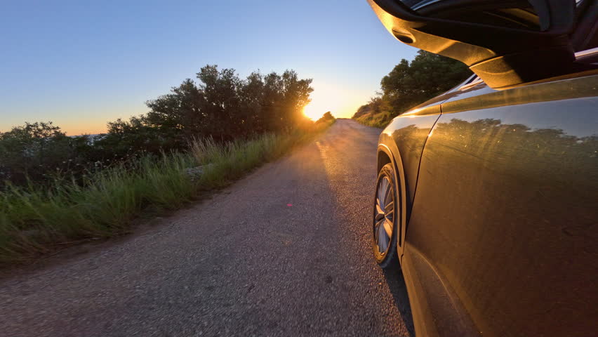 LENS FLARE, LOW ANGLE VIEW: Black car travels along a coastal asphalt road with a view of golden sun slowly setting over the blue Adriatic Sea. Picturesque summer road trips on sunny island of Hvar.