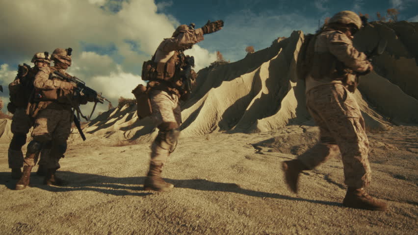 Four Soldiers in Desert Camouflage Uniforms and Tactical Gear Walk in Formation Across a Rocky, Sandy Middle Eastern Landscape Holding Rifles and Moving Cautiously on a Mission. Special Ops Operation