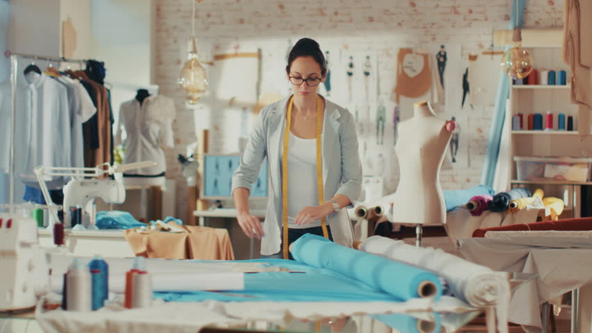 Beautiful Female Fashion Designer Adjusting Fabrics on a Tailored Mannequin. In Her Studio Various Sewing Items and Colorful Fabrics Laying around, Mannequins Standing, and Sketches Pinned.