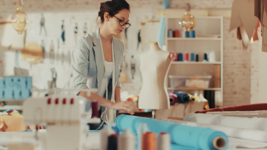 Beautiful Female Fashion Designer Adjusting Fabrics on a Tailored Mannequin. In Her Studio Various Sewing Items and Colorful Fabrics Laying around, Mannequins Standing, and Sketches Pinned.