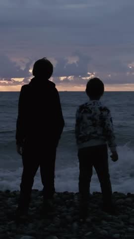 Two boys standing on the rocks of a pebble beach on the ocean shore and throwing stones into the water. Silhouettes of teenagers at sunset by the sea in the evening against background of sunset sky