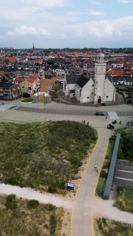 Aerial view of a coastal European town with red-roofed houses, a central church, and a geometric garden. A wide road with cars and a bike lane runs along the foreground near sand dunes.