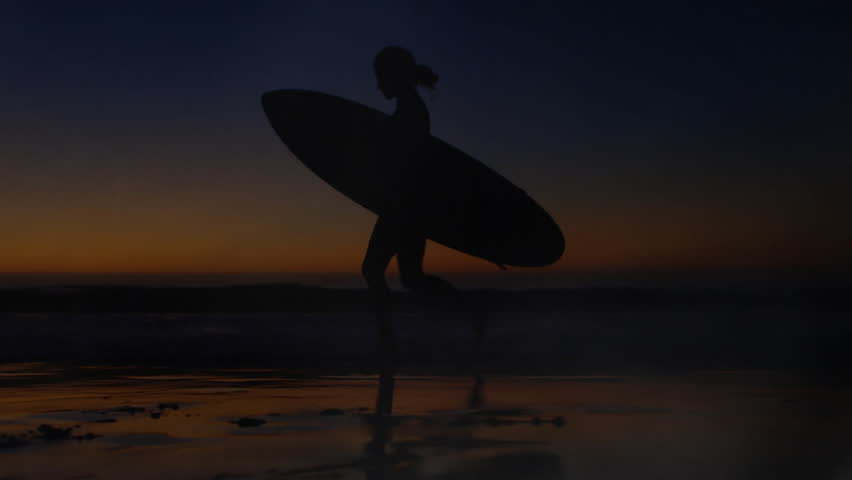 Female surfer emerging from shoreline walking across wet sand at dusk swirling mist for surf promo. Silhouette, coastal, sunset, mist, shoreline, ocean, tranquility