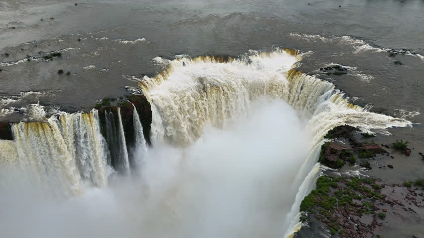 Cascades of Iguazu Falls drop into swirling river below with green rainforest on cliffs. The aerial view captures energy and movement. Water roars between rocks filling the deep canyon with spray