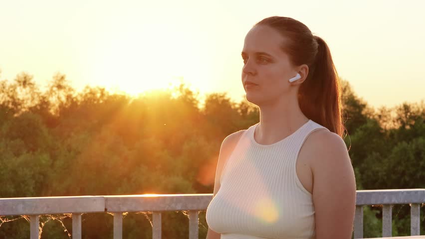 A young woman in a white tank top with a ponytail stretches her arms, one across her body, as the sun sets behind her outdoors.