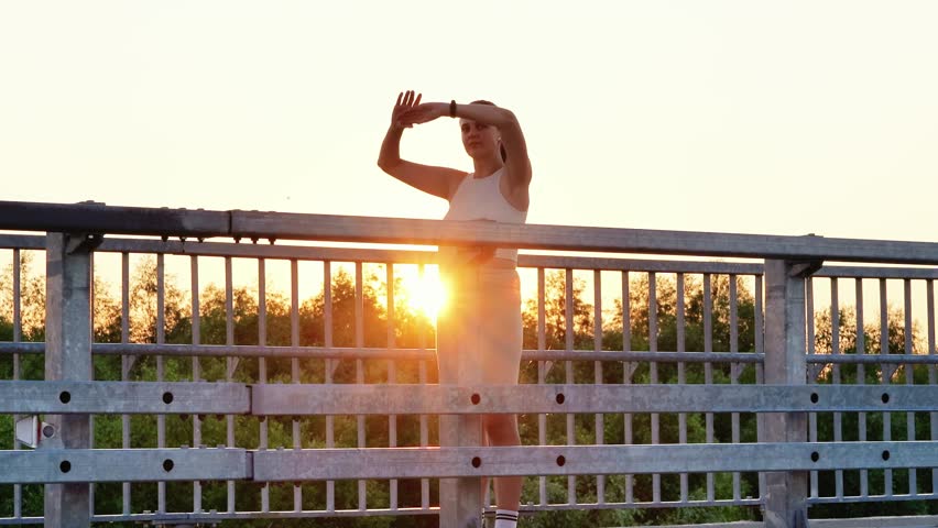 A young woman in a white tank top and light-colored shorts stretches her arms overhead on a bridge, silhouetted by the setting sun.