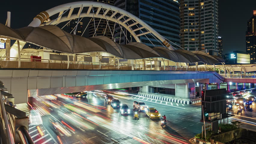 Bangkok timelapse stunning view of a modern pedestrian bridge illuminated against the night sky. Below busy traffic flows with cars and motorcycles. The urban setting exudes a vibrant nightlife