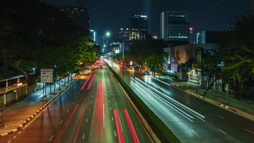 bangkok timelapse Aerial view of a bustling urban street at night showcasing light trails from moving vehicles. Lush greenery line the road illuminating the city skyline in the background