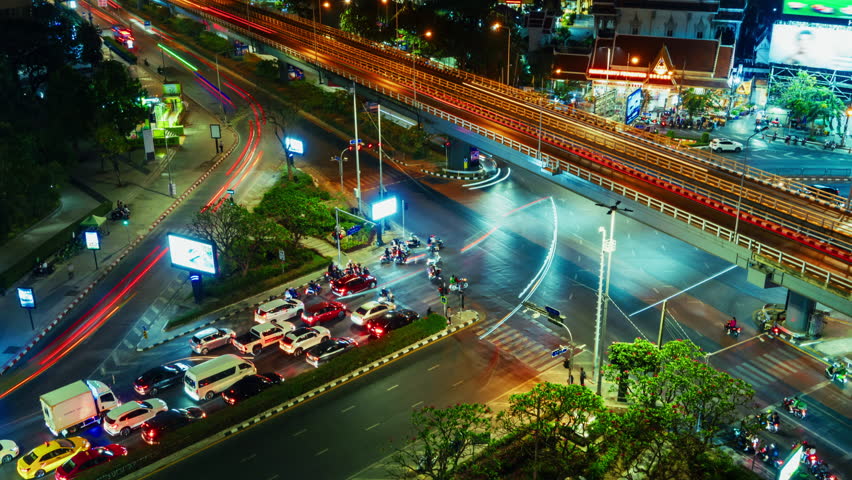 Bangkok timelapse Aerial view of nighttime urban traffic showcasing vibrant light trails from moving vehicles along multi-lane roads and a motorway overhead. Illuminated billboards and streetlights 