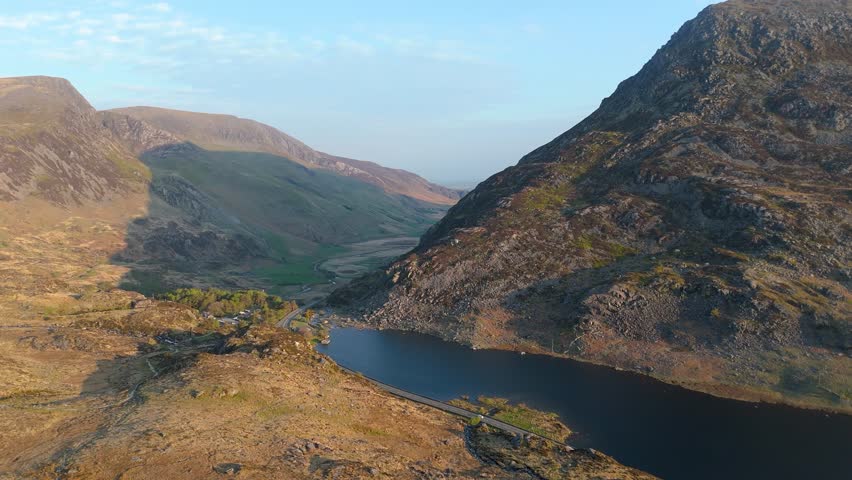 A drone scenery of Llyn Ogwen lake with A5 road along the shoreline between mountains in Snowdonia National Park, Wales during morning with blue sky