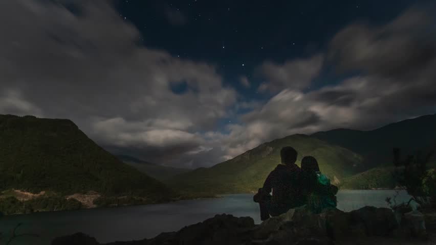 A Couple silhouettes sit beside a mountain lake under a time lapse starry night sky with drifting moonlit clouds