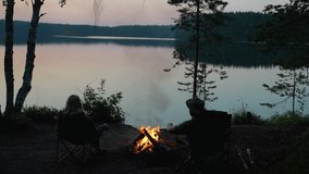 A man and a woman travelers sitting by a campfire on the shore of a forest lake, enjoying the sunset. A couple warming themselves by the campfire, enjoying the view of the lake. Tourists by the - Powered by Shutterstock - Get 15% off with code: PIKWIZARD15