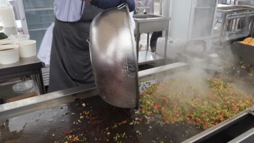 A chef works on a flat top grill, dicing and stirring minced meat with one spatula while another spatula is used to add and mix in a large pile of finely chopped vegetables.