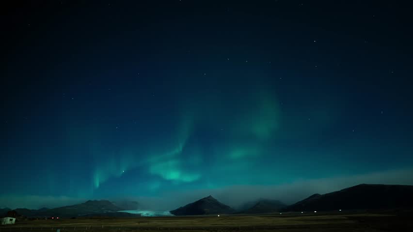 Timelapse of aurora borealis swirling through a starry, partly cloudy sky above black mountain silhouettes