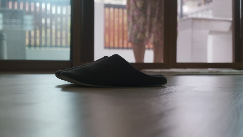 Black slipper on wooden floor with old lady's bare foot stepping indoor near soft light at home