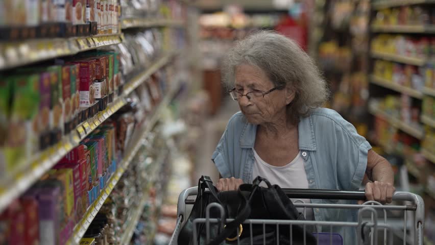 An elderly senior woman navigates a busy grocery store aisle, carefully examining the shelves stocked with various food items as she pushes her shopping cart. - Powered by Shutterstock - Get 15% off with code: PIKWIZARD15