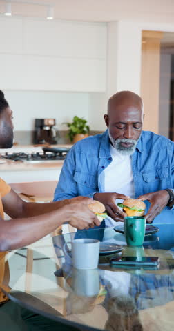 Vertical video: African American father and son passing sandwich, eating while chatting at home. Family, bonding, breakfast, communication, casual, home, vertical video