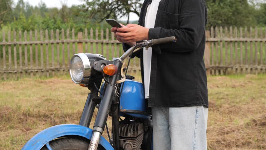 Farmer using smartphone next to vintage motorcycle in countryside
