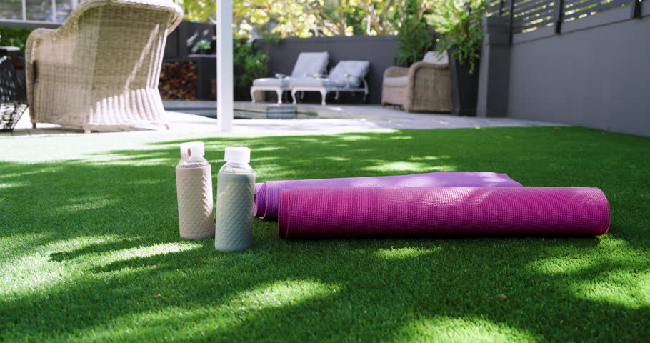 Camera dollying forward showcasing purple yoga mat with two water bottles on backyard patio turf. Outdoor, wellness, relaxation, leisure, modern, natural, vibrant