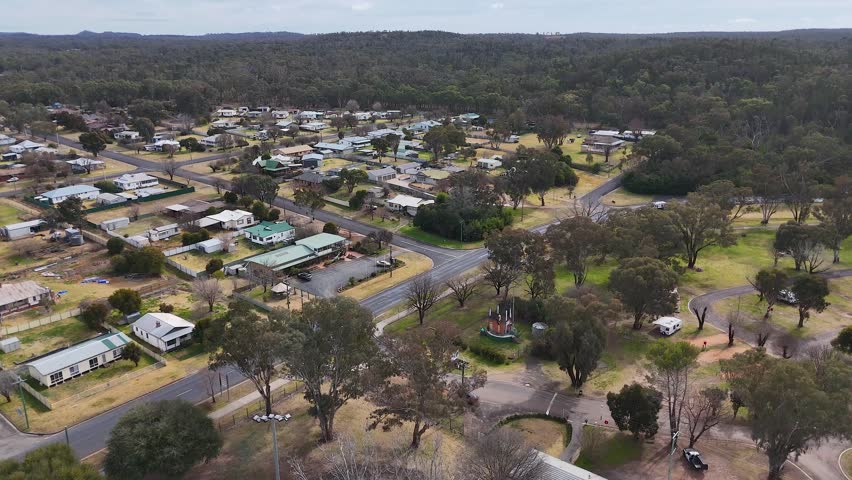 Drone footage glides above a quiet rural neighborhood, highlighting local streets, houses, and open green spaces under overcast natural daylight