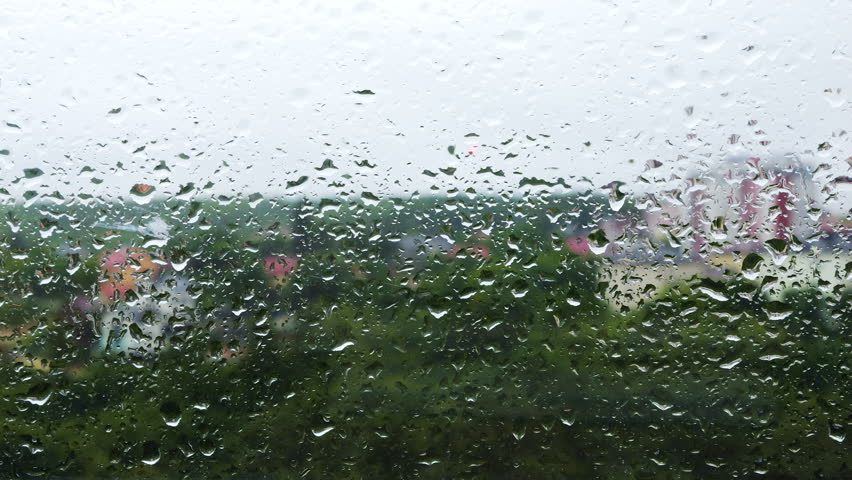 Water drops roll down the window glass during rain against the background of forest trees and city buildings.