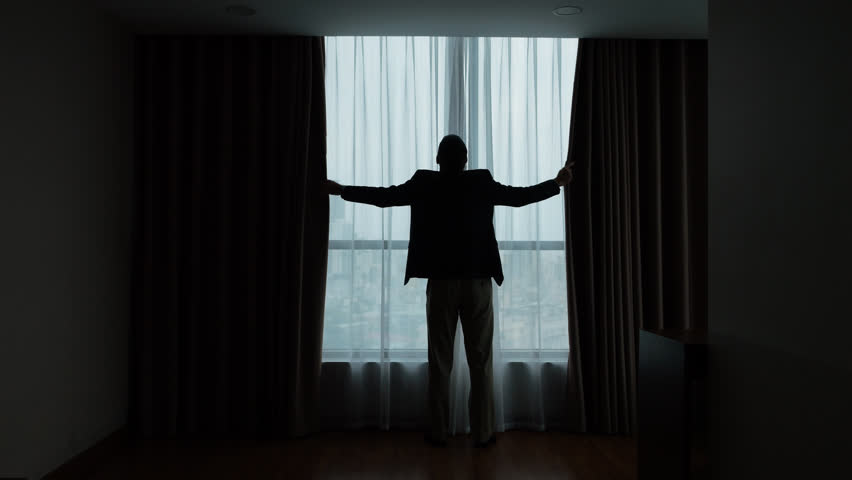 Man stands by the window, using both hands to close heavy blackout curtains, darkening the room almost completely. Slow motion, full-length silhouetted shot from behind.