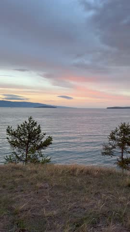 Vertical video of summer evening on Lake Baikal. Panoramic view of Olkhon Island coast and pink clouds at sunset. Natural background. Picturesque sunset landscape