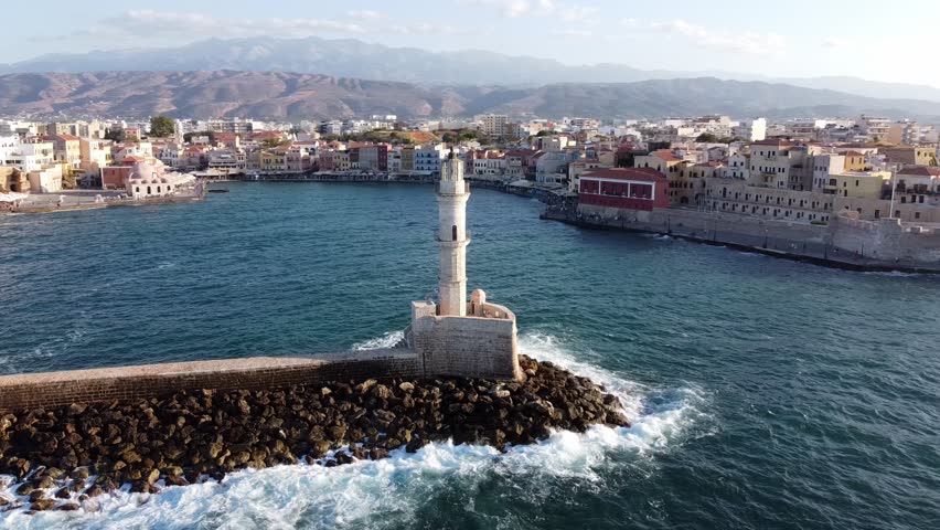 Drone circles around lighthouse in old Venetian harbor of Chania, Crete, historic town and mountains in background, Mediterranean sea
