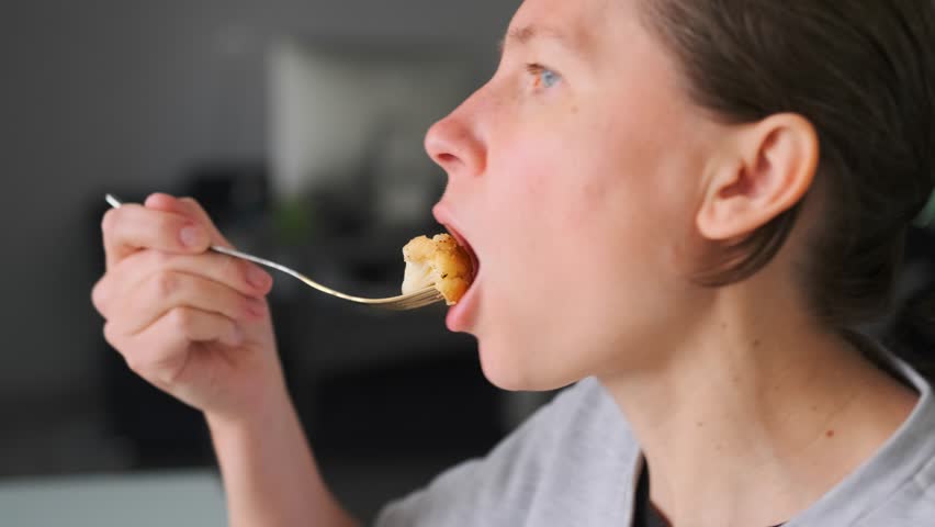 Close-up profile of a woman blowing on a hot bite of homemade fried cauliflower before tasting it, chews slowly, enjoying the moment. Pleasure of eating simple food at home.