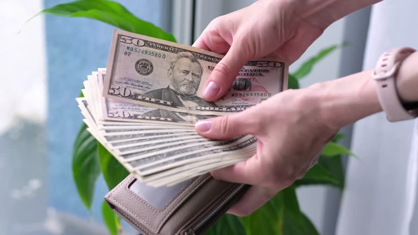 A person is holding a wallet with a lot of money in it. The wallet is brown and has a clear window