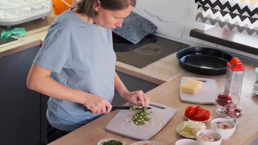 Female slicing fresh green onions on a cutting board while filming a healthy homemade pizza recipe. The countertop features natural ingredients like tomatoes, cheese, and herbs. Cooking content.