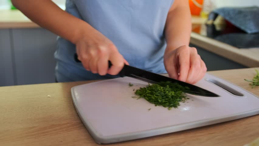 Woman slices green onions in clean, well-lit kitchen, filming a vlog about healthy homemade pizza. The cutting board is surrounded by fresh ingredients like tomatoes, cheese, and herbs, home cooking.