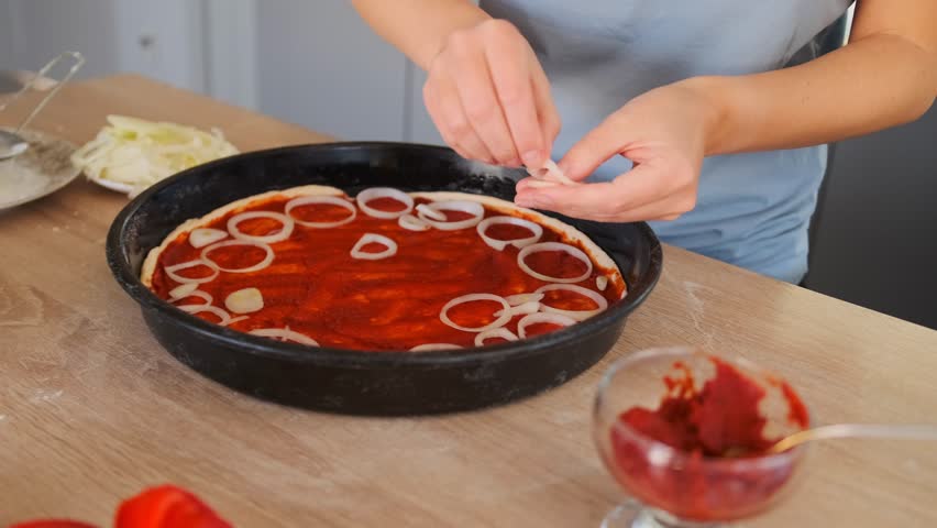 Close-up top view of female hands skillfully placing raw onion rings on pizza dough already covered with tomato paste. Pizza lies on a wooden table surrounded by ingredients, cozy homemade cooking.