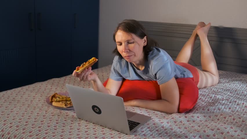 Close-up, woman with short hair lying on her stomach in bed, biting slice of pizza while typing messages on laptop. She chatting casually with friends on social media. Cozy and relaxed home evening.