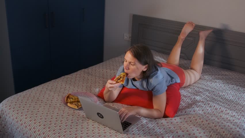 Young woman with short hair lies on her stomach in bed, typing messages on a laptop while taking a bite of pizza. Close-up of a peaceful evening alone, combining digital communication, dining at home.