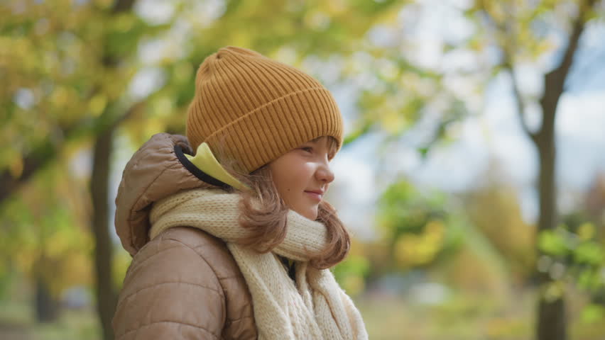 close up of cute girl standing while holding yellow autumn leaf in hand wearing mustard beanie beige quilted jacket and knitted scarf under golden tree canopy on leaf strewn