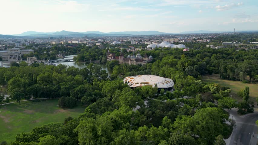 Aerial view of the Vajdahunyad Castle and surrounding lush green trees, contrasting with the city skyline in the background, Budapest, Hungary.