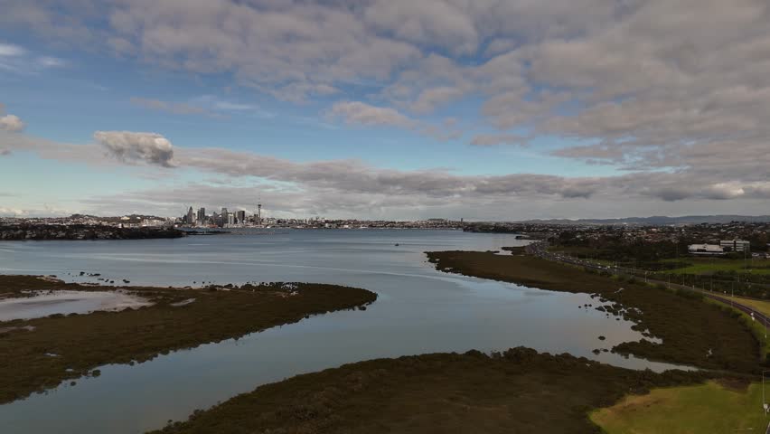 A scenic 4K 60fps aerial flight over the tranquil waters of Shoal Bay, revealing the stunning Auckland CBD skyline, including the Sky Tower, in the background. A perfect shot of NZ city life
