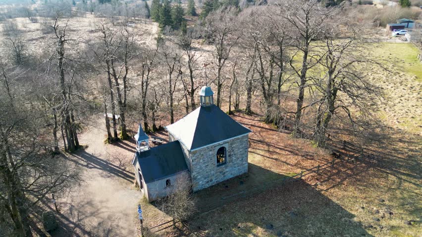 Aerial view of a stone chapel with a dark roof, surrounded by bare trees casting long shadows on the grassy ground, Malmedy, Wallonia, Belgium.