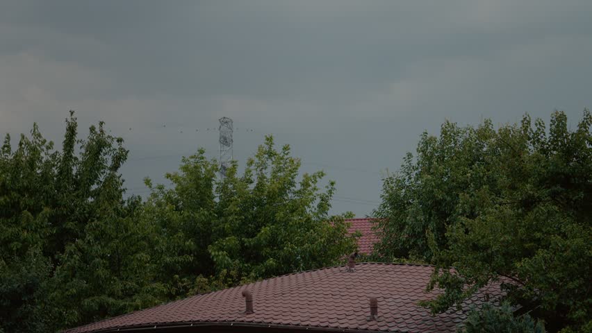 Dark clouds loom over the horizon as lightning flashes in the blue sky near high voltage wires and trees, signaling the arrival of a powerful storm.