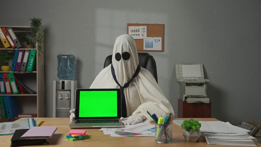 Person dressed as ghost day at work. Halloween businessman ghost in white sheet sits at office desk with laptop chroma key green screen, pointing shows thumb up. Happy halloween business concept.