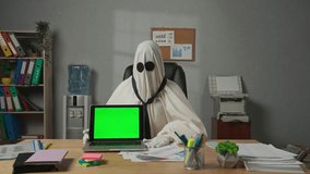 Person dressed as ghost day at work. Halloween businessman ghost in white sheet sits at office desk with laptop chroma key green screen, pointing shows thumb up. Happy halloween business concept. - Powered by Shutterstock - Get 15% off with code: PIKWIZARD15
