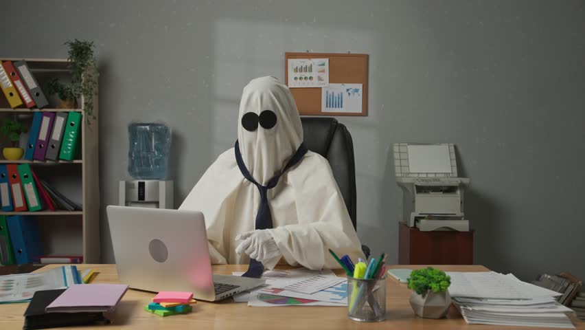 Person dressed as ghost day at work. Halloween businessman ghost in white sheet sits at office desk works on laptop, sad expression disappointed shrugs shaking head. Happy halloween business concept.