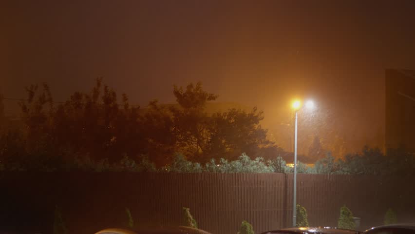 Strong winds and heavy rain batter homes and parked cars, creating a dramatic atmosphere under dark clouds with occasional flashes of lightning illuminating the scene.