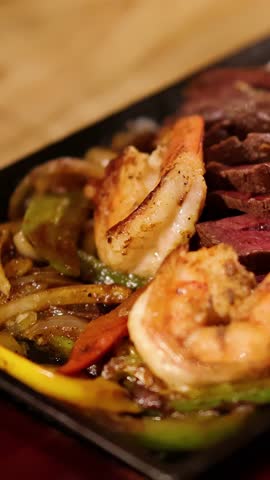 A close-up view of steak and shrimp fajitas on a hot skillet, with a hand slicing steak under warm, ambient restaurant lighting. Shallow depth of field
