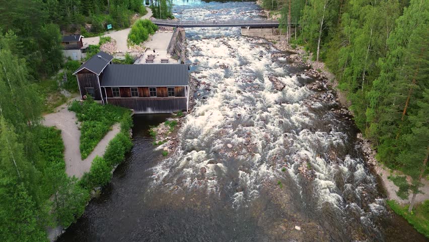 The rapids roar between the rocks in the Finnish landscape, with water flowing powerfully as a stream between strips of forest. An old wooden bridge spans above the rapids.