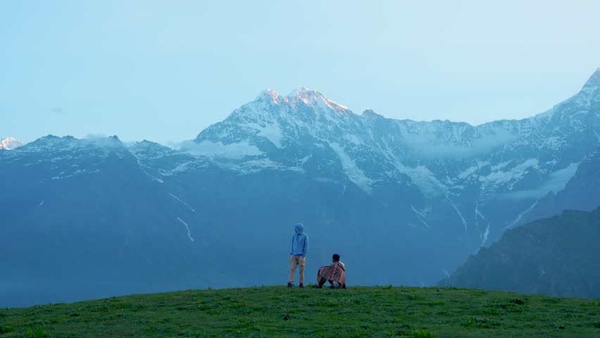 4K video of two men at the Buda Madhyamaheshwar Viewpoint on top of a mountain at sunrise, with a clear view of the Chaukhamba peaks in Uttarakhand, India.	