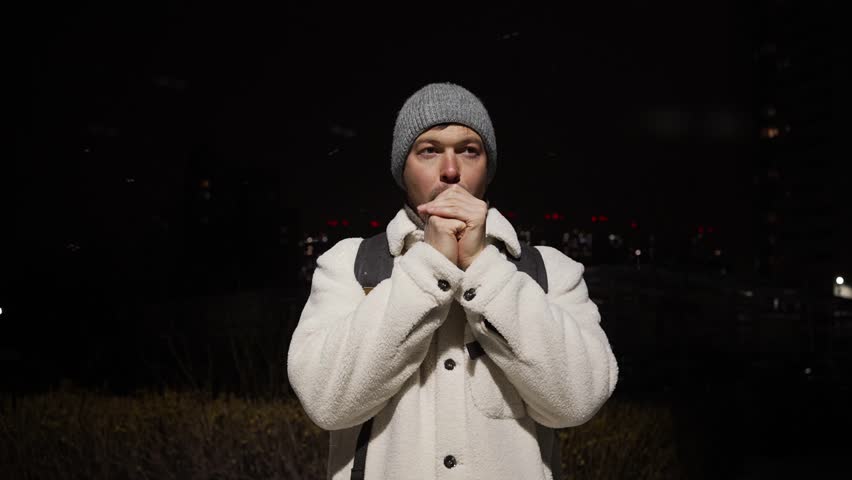 Cold traveler enduring freezing cityscape, huddling in thick layers, warming numb hands through desperate breathing and friction against stark winter environment
