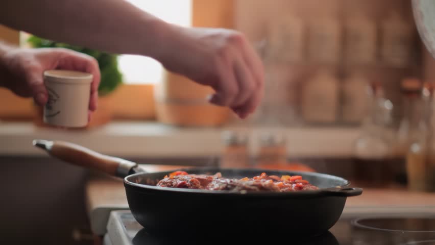 A man's hands sprinkle spices into a hot, steaming pan where an Osso Buco steak is simmering. Spices and herbs are blurred in the background. Gourmet home cooking in progress