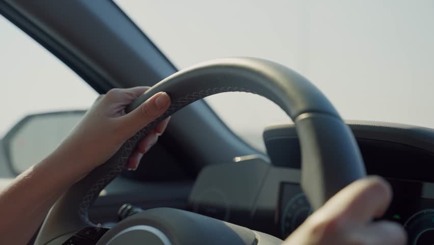 Female hands holding steering wheel driving vehicle or modern sedan car in highway. Car ride as woman commuting to work or going for vacation on sunny day sun shining into dashboard as cars pass by
